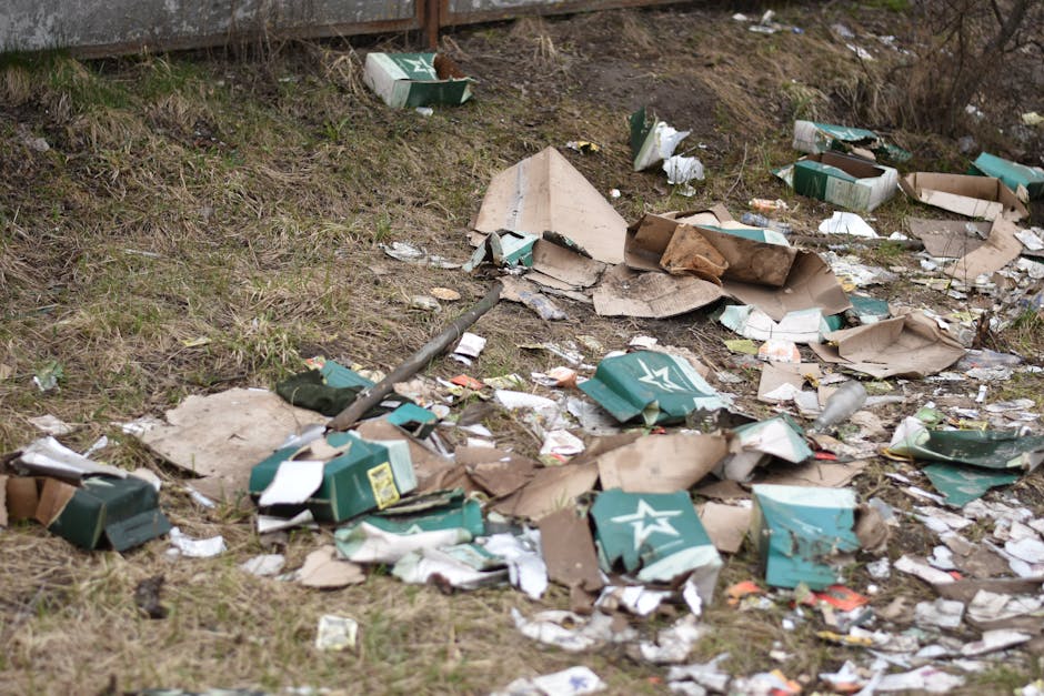 A patch of ground next to a curb or pathway is littered with scattered rubbish, predominantly consisting of flattened cardboard boxes with green and white branding, some crumpled and torn. Alongside t