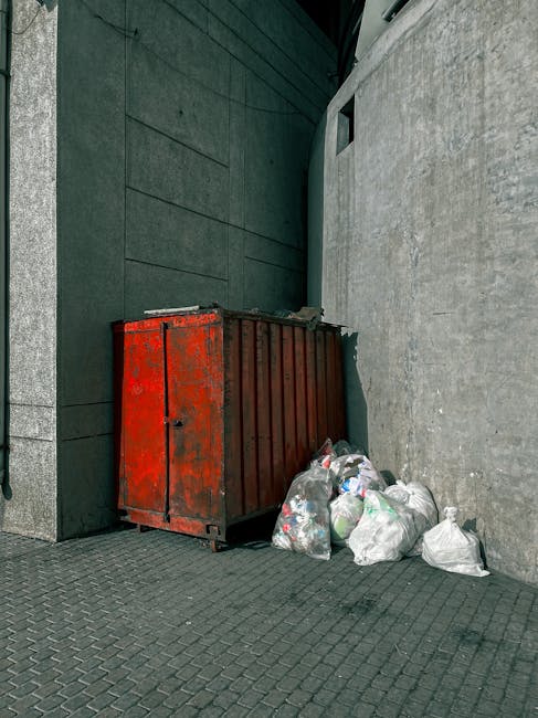 A large collection of overflowing waste bins and numerous discarded rubbish bags and cardboard boxes piled on a paved sidewalk in front of a commercial building. The rubbish includes black, red, and g