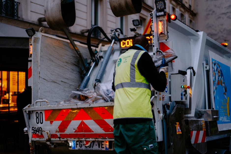 A waste management worker, dressed in an orange high-visibility uniform with reflective yellow and silver stripes, is seen from above standing on a sidewalk next to a concrete wall, holding a long-han